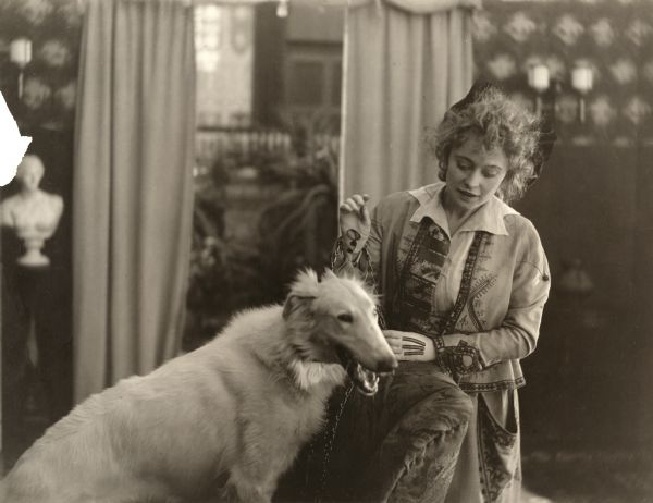 Actress Jackie Saunders plays with a white Russian wolfhound in a scene still for the silent drama "A Bit of Kindling." Saunders wears an interesting folk pattern jacket and skirt with a blouse that has a star-shaped collar.