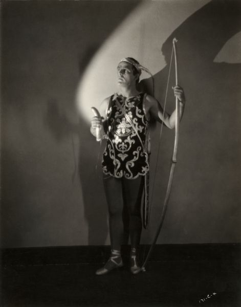 In this dramatically lit publicity still for the silent drama "Look Your Best," Antonio Moreno wears what appears to be a cupid costume with feathered headband, bow, quiver, and dancing shoes. He holds a large false nose in his hand. Carlo Bruni, Moreno's character in the film, is a dancer.