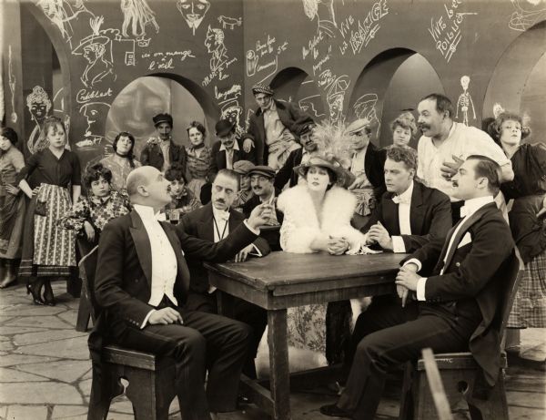 In an Apache dance hall with artists' sketches chalked upon the walls, the opera singer Sonia Smirnov (played by Olga Petrova) sits at a rough table surrounded by her male admirers. To her right is Lachaise (played by Count Carl Gustaf Sixtensson Lewenhaupt), and on her left is Alan Hall (Mahlon Hamilton) in this scene still for the silent drama "The Black Butterfly." A dozen or more unidentified actors costumed as members of the Parisian underworld are in the background.