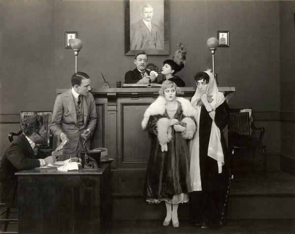 In this courtroom scene still for the Keystone silent comedy "Black Eyes and Blue," the judge is played by Jack Henderson. The woman in the black hat is probably his wife, played by Dorothy Hagan, and the blonde woman is Stella, played by Juanita Hansen. The other actors are unidentified.