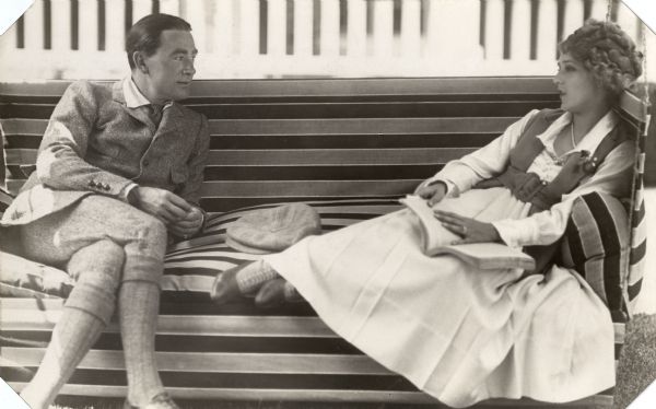 Silent film couple Owen Moore and Mary Pickford sit on a porch swing. He wears a herringbone worsted wool suit with large patch pockets, and plus fours. She wears a loose jacket with a wide belt over a white pleated dress and has a book in her hands.