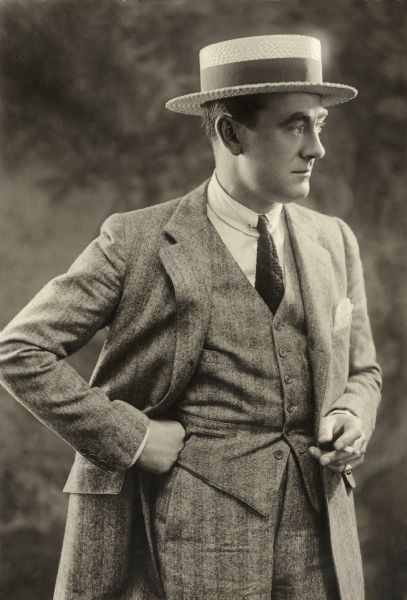 Silent film star Owen Moore stands holding a cigarette in a half length studio portrait. He wears a herringbone worsted wool suit, white shirt with soft collar and collar bar, a knit necktie, and a straw boater hat.