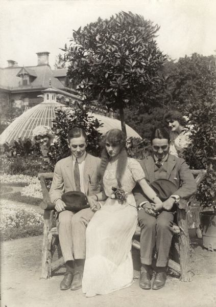 Silent film actors Matt Moore (left) and his brother Owen Moore sit on either side of Florence Lawrence who holds both their hands on a rustic bench in the garden of a wealthy home. Behind the bushes, two unidentified actresses spy on the love triangle in this scene still from a Victor Film Company production from 1912.