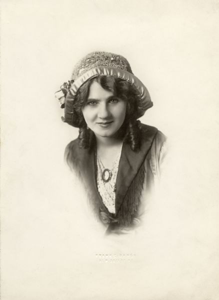 Head and shoulders studio portrait of Florence Lawrence wearing a hat. She wears a cameo and looks directly at the camera.