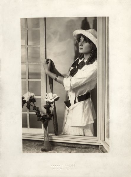 Florence Lawrence poses in an elaborate set in the studio of portrait photographer Frank C. Bangs in New York. She stands looking in an open window with a tennis racquet in her hands. A vase with roses is in the foreground.