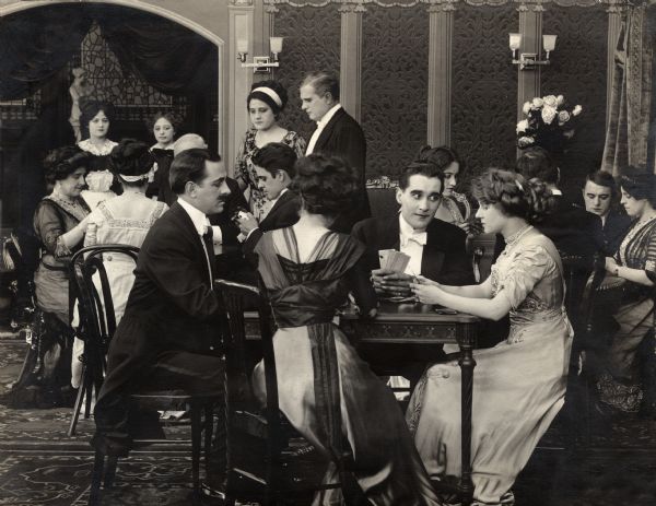 In a scene still for the 1911 Lubin silent drama "The Snare of Society," Florence Lawrence, right foreground in the light-colored dress, is a society woman who has become addicted to gambling. The man who looks at her so intently is Jack Standing. The other actors in the elegant gambling salon are unidentified.