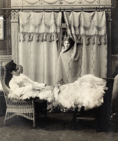 Florence Lawrence peeps through curtains to look at Arthur V. Johnson sleeping in a wicker chair under large white furs in a scene still for the 1911 silent drama "One on Reno."