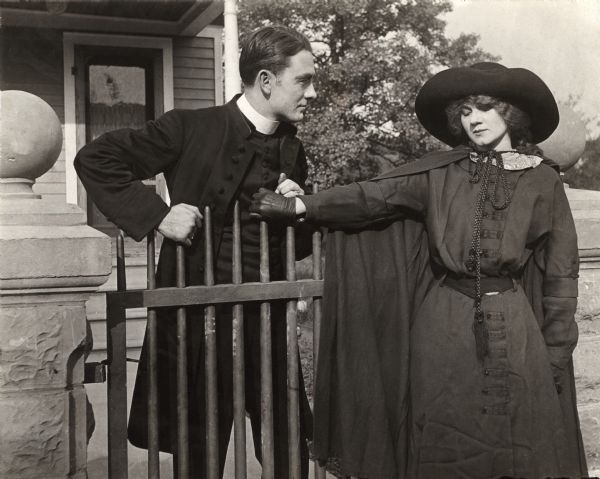 Owen Moore, costumed as a minister, talks to Florence Lawrence by a gate in front of a house in a scene still from a silent drama, possibly "The Redemption of Riverton" from 1912.