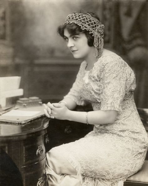 Lubin leading lady Lottie Briscoe sits at a writing desk in a publicity still. She wears a white lace overdress and a beaded Juliet cap.