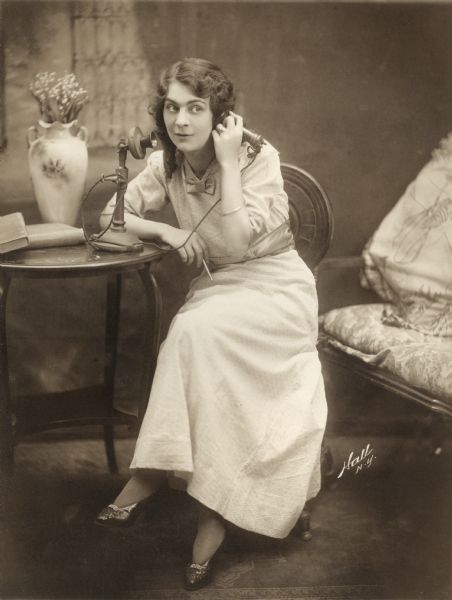 Silent film actress Lottie Briscoe in a studio publicity photograph posed sitting at a small table with a candlestick telephone, a vase with flowers, and some books.