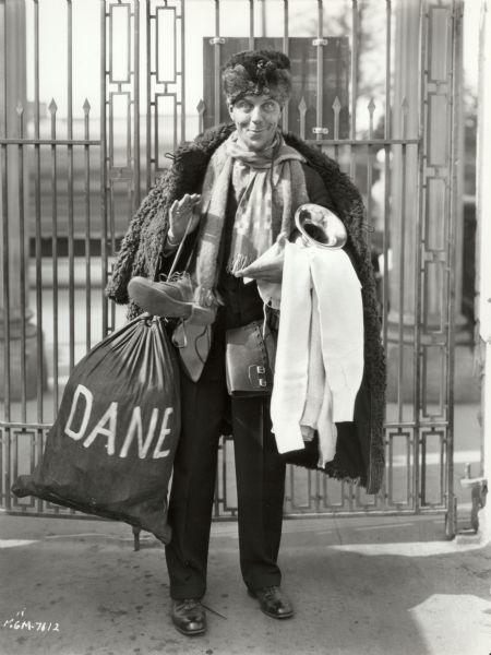 Karl Dane wearing and carrying a variety of clothes, including a fur hat, poses by the MGM studio gate in a 1927 publicity still.