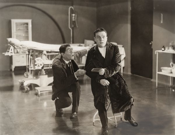 In a medical operating room, Robert Sandell (played by Raymond McKee) is shocked by what he has read in the notepad that the Hunchback (Lon Chaney) has given him in a scene still for the silent horror film "A Blind Bargain."