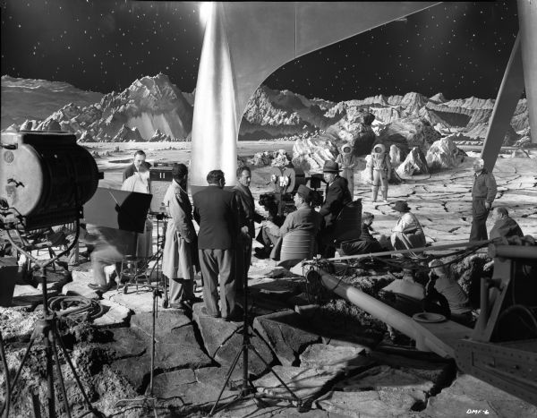 Cast and crew of the 1950 science fiction film "Destination Moon" are on a lunar set including one leg of a gigantic spacecraft. Facing out, Lionel Lindon, the cinematographer, is having a discussion with a man seated behind the large Technicolor camera on a crane. Two astronauts are in the background on the moon's surface.