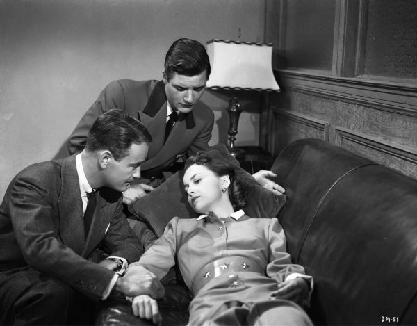 In a scene still for the 1946 film "The Dark Mirror," Olivia de Haviland lies on a couch looking worried as Lew Aires holds her wrist and Richard Long watches with concern.
