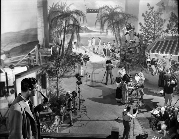 Robert Cummings, left foreground with sunglasses in his hand, looks down upon a busy advertising photography studio in 1948 romantic comedy "Let's Live a Little." Three stills cameras are visible: a 4x5 press camera on a tripod in front of a man in an easy chair (lower right), an 8x10 view camera with dark cloth pointing toward the travel trailer set on the right, and another 8x10 camera with a photographer directing women in a beach scene toward the back.