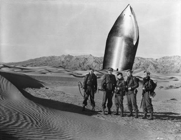 The crew of Rocketship X-M survey the rugged terrain of Mars. Behind them is their wrecked ship. From left to right they are played by Noah Beery Jr., John Emery, Osa Massen, Lloyd Bridges,  and Hugh O'Brian.