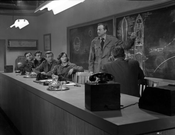A scene still for "Rocketship X-M" with, from left to right, Noah Beery Jr., Hugh O'Brian, Lloyd Bridges, Osa Massen, and John Emery in a conference room. John Emery stands and points to a diagram of the rocketship drawn on a blackboard.