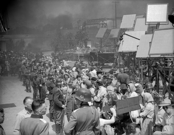 A large crowd of extras costumed as Ukrainian villagers stands guarded by men in World War II German uniforms in a production still for "The North Star." In the background is smoke from the burning village set. In the foreground is a motion picture camera, and in the upper right corner are ten or more large reflectors.