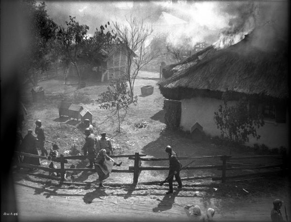 In this high angle shot, Ukrainian villagers and German soldiers flee a burning village in a production still for "The North Star." In the foreground a woman in peasant costume climbs through a split-rail fence. Behind her is a farmyard with chicken houses. Beyond that are farm buildings and houses with burning thatched roofs.