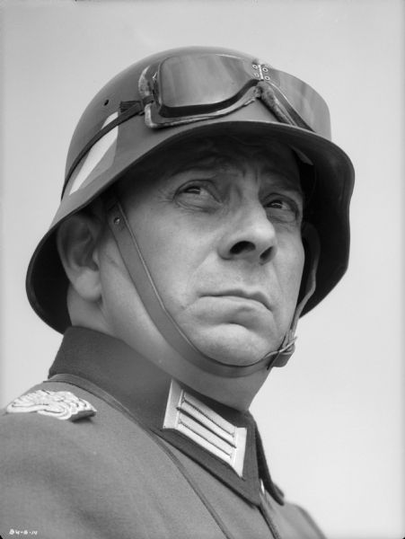 Erich von Stroheim poses with his right shoulder toward the camera in his costume as the German military doctor Otto von Harden in a publicity still for "The North Star." He wears a steel helmet (Stahlhelm) on which are stored a pair of goggles.