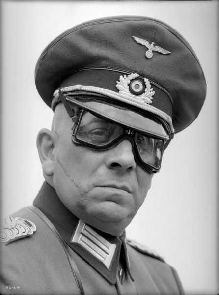 Erich von Stroheim poses with his right shoulder toward the camera in his costume as the German military doctor Otto von Harden in a publicity still for "The North Star." An officer's visor cap is on his head, and goggles cover his eyes. A prominent scar runs across his right cheek.