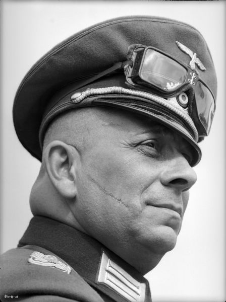 Erich von Stroheim poses with his right shoulder toward the camera in his costume as the German military doctor Otto von Harden in a publicity still for "The North Star." An officer's visor cap is on his head with goggles stored above the visor. A prominent scar runs across his right cheek, and he is faintly smiling.