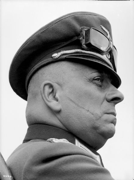 Erich von Stroheim poses in right profile in his costume as the German military doctor Otto von Harden in a publicity still for "The North Star." An officer's visor cap is on his head with goggles stored above the visor. A prominent scar runs across his right cheek.