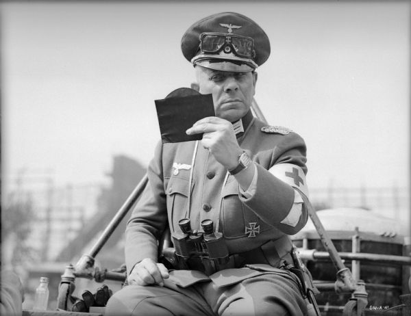 With a critical eye, Erich von Stroheim examines his make-up in a hand mirror. He sits outdoors in costume as the German military doctor Otto von Harden in a publicity still for "The North Star." An officer's visor cap is on his head with goggles stored above the visor, binoculars hang from a strap on his neck, and a Red Cross armband is on his left arm.