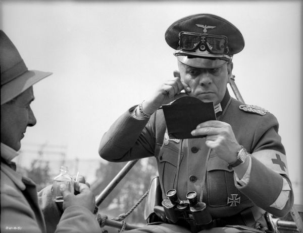 At left, the make-up artist, probably Robert Stephanoff, watches as Erich von Stroheim pinches his right cheek and examines his make-up in a hand mirror. Von Stroheim is sitting outdoors in costume as the German military doctor Otto von Harden in a publicity still for "The North Star." An officer's visor cap is on his head with goggles stored above the visor, binoculars hang from a strap on his neck, and a Red Cross armband is on his left arm.