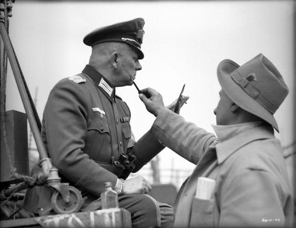 Erich von Stroheim watches in a hand mirror as a make-up artist, probably Robert Stephanoff, colors a scar on his cheek with a pencil. Von Stroheim is sitting outdoors in costume as the German military doctor Otto von Harden in a publicity still for "The North Star."