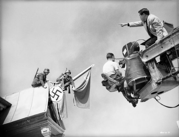 Cinematographer James Wong Howe high up on a camera crane directs actors playing German soldiers who are replacing the Soviet flag flying from a flagstaff on a roof gable with the German battle flag.