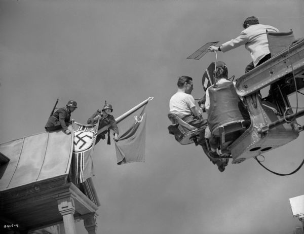 High up on a camera crane, cinematographer James Wong Howe adjusts a flag over the cameraman's head. In front of the camera, two actors playing German soldiers replace the Soviet Flag flying from a flagstaff on a roof gable with the German battle flag.