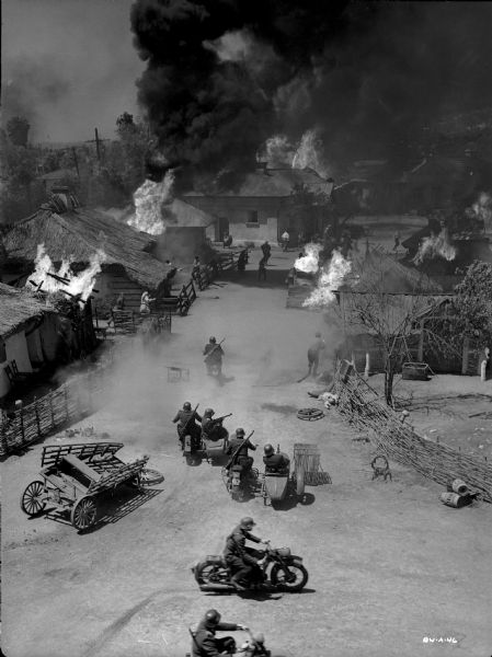 In this high angle shot, Ukrainians are setting fire to their own village as German soldiers arrive on motorcycles, some with sidecars, in a production still for "The North Star."