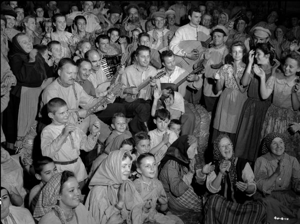 Scores of actors costumed as Ukrainian village men, women, and children clap and sing in a production still for "The North Star." Six musicians standing in the middle of the crowd play the accordion, domra, and several sizes of balalaika.