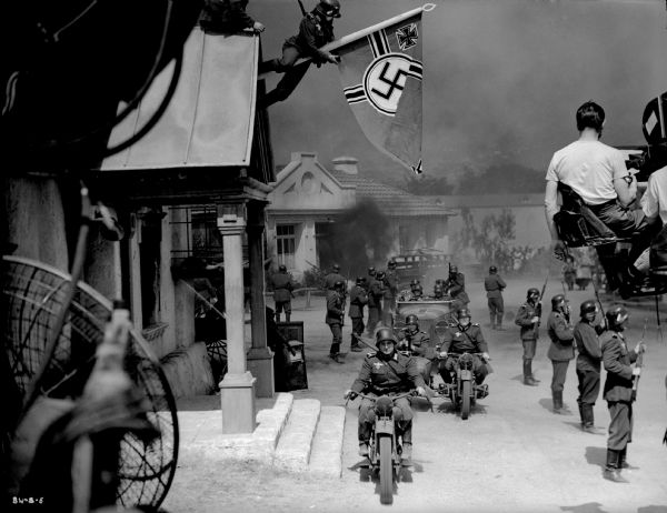 A line of German foot soldiers and an automobile with officers and their motorcycle escort entering a Ukrainian village in a production still for "The North Star." A soldier on the gable of a building has hung a German battle flag. Motion picture equipment is in the foreground, on the left a large fan, on the right a camera and its crew on a crane.