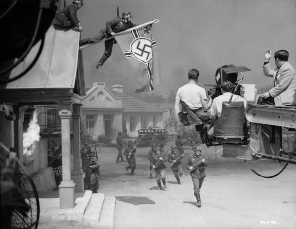 Five German foot soldiers run toward the camera with bayonets on their rifles while another shoots water on a burning building on the left in a production still for "The North Star." A soldier climbs out on a staff attached to the gable of a building to hang a German battle flag. Motion picture equipment is in the foreground, on the left a large fan, on the right a camera and its crew on a crane. The cinematographer James Wong Howe on the crane at the far right seems to direct the action