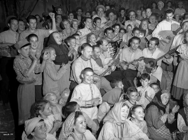Scores of actors costumed as Ukrainian village men, women, and children clap and sing in a production still for "The North Star." Six musicians standing in the middle of the crowd play the accordion, domra, and several sizes of balalaika.