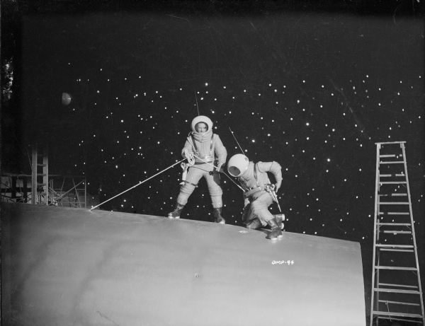 Two actors in space suits and suction cup boots work on the exterior of a spaceship in a production still for "Destination Moon." Behind them is a black sky filled with stars. On either side is production equipment, a step ladder and scaffolding.