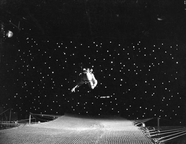 An actor in a space suit "floats" in front of a background of stars. Below him is a safety net and a technician watching on the right.