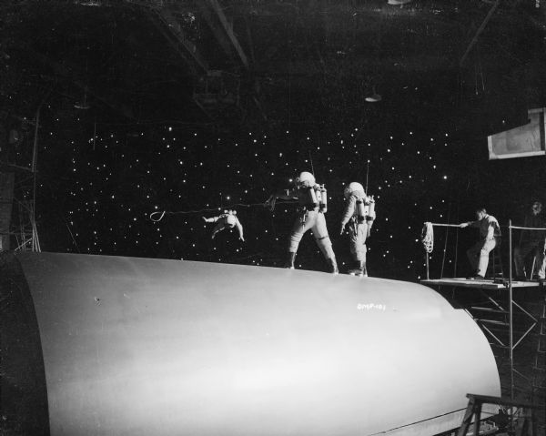 In a production still for "Destination Moon," an astronaut in a space suit is floating away from the spaceship. Two of his companions stand on the hull of the ship, one of whom attempts to lasso the lost astronaut. On the right, a technician on a scaffold watches the scene.