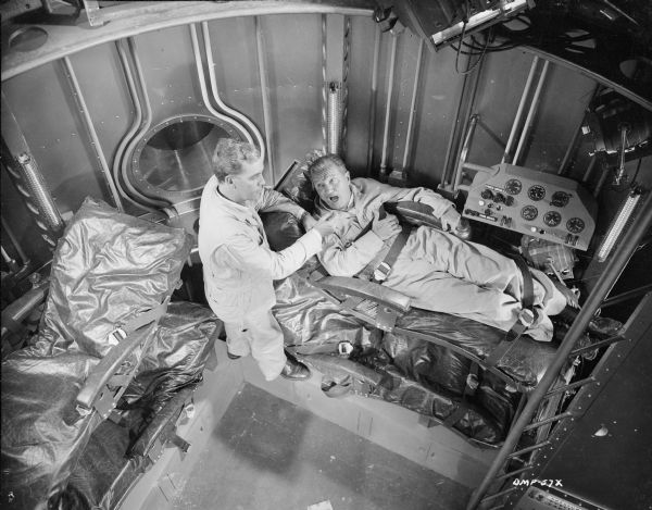 Actors John Archer (left) and Tom Powers (right) pose in the set for the interior of a spacecraft. Powers is buckled into a padded bunk.