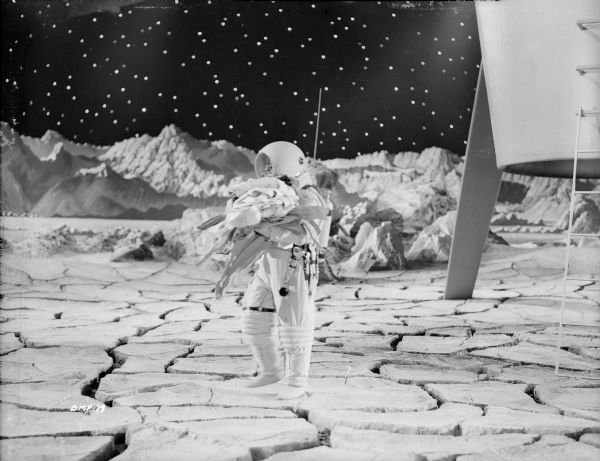 An astronaut in a spacesuit carries some stiff laundry on the surface of the moon in scene still for "Destination Moon." On his belt are a flashlight, an adjustable wrench, and other tools. Behind him is one leg of the spaceship and its ladder.