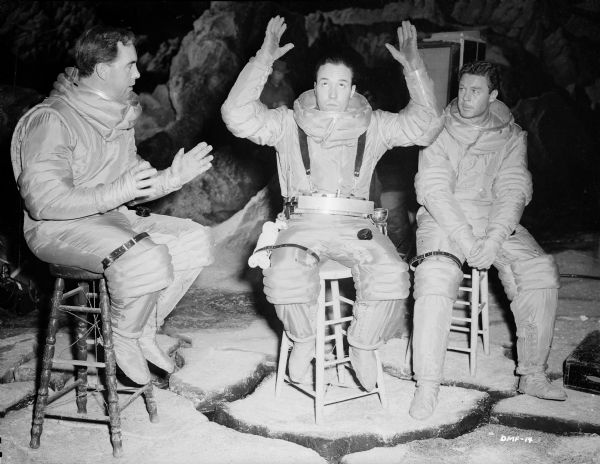 From left to right, the actors Warner Anderson, Dick Wesson, and John Archer relax on a lunar set during the filming of "Destination Moon." They sit on stools and wear spacesuit costumes without helmets.