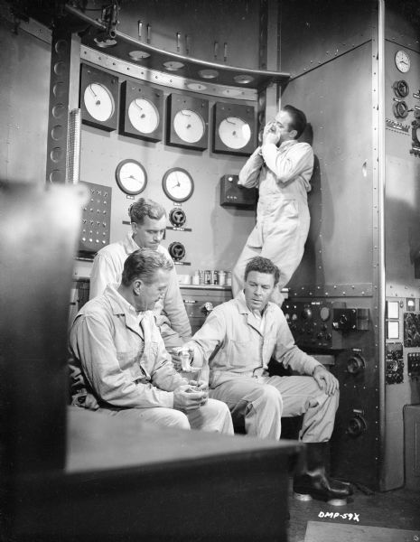 Counter-clockwise, the actors Warner Anderson, Tom Powers, John Archer, and Dick Wesson (playing the harmonica) are in the control room of a spaceship. Archer holds out an empty beverage container to Powers.