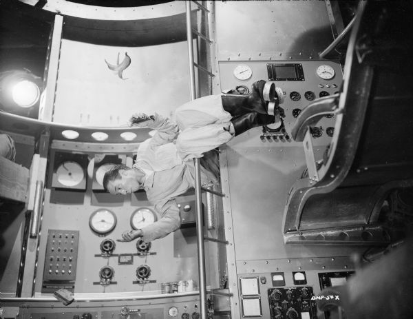 In a demonstration of weightlessness, Dick Wesson sits sideways on a ladder in the interior of a spaceship. Floating near him, hung by threads, are a sandwich and a banana.