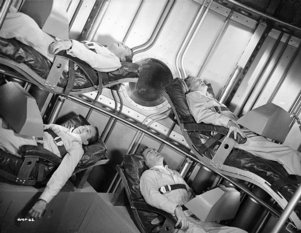 Photographed at an angle, four astronauts are strapped into their bunks in a spaceship interior. From top left, proceeding clockwise, the actors are John Archer, Tom Powers (grimacing), Warner Anderson, and Dick Wesson. All appear to be straining. Only Wesson has his eyes open.