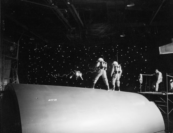 Two astronauts stand on the hull of a spaceship offering a line to a third astronaut who floats against a background of stars. On the right, a technician on a scaffold watches the scene.