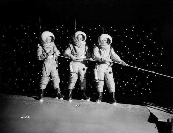 Three astronauts stand on the hull of a spaceship holding a rope extending out of the frame. Behind them is the black of space and bright stars.