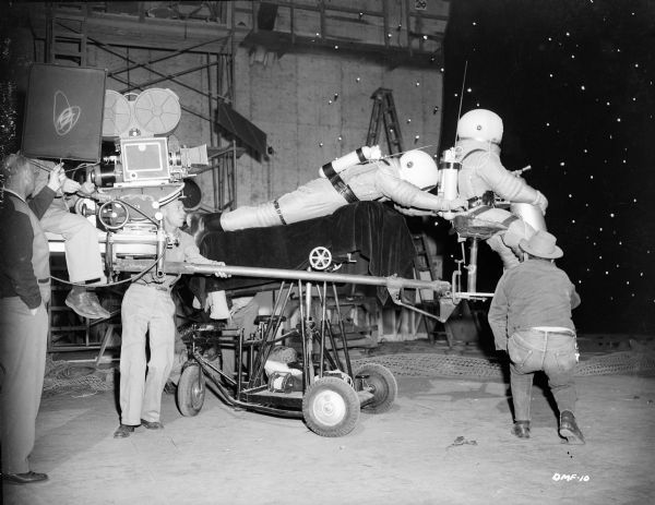 An interesting production still showing how the rescue of a spacewalking astronaut was filmed. One astronaut, holding a gas canister for propulsion, sits on chair mounted to the front of the Technicolor camera's crane. The astronaut who catches him lies on his stomach on a dolly covered with black velvet. Crouching low, various technicians maneuver the actors against a background of stars.