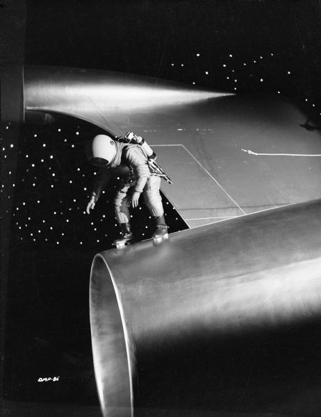 Standing at the extreme tail end of a spaceship, near the rocket's exhaust, an astronaut bends down. His disconnected safety line floats behind him.